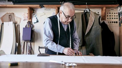 Older man with a tape measure around his neck working as a tailor, with patterns and fabric pieces hanging on the wall behind